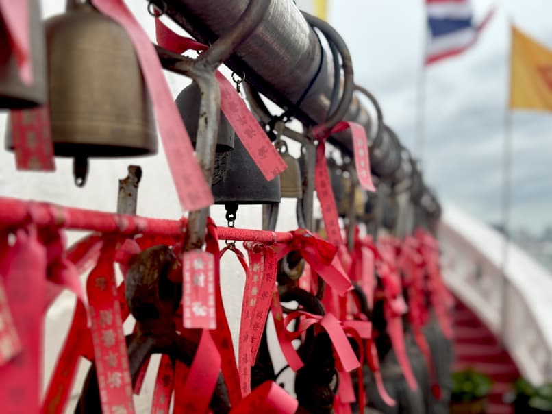 Prayer bells with red ribbons and tags hanging at a temple, with Thai and buddhist flags in the background.